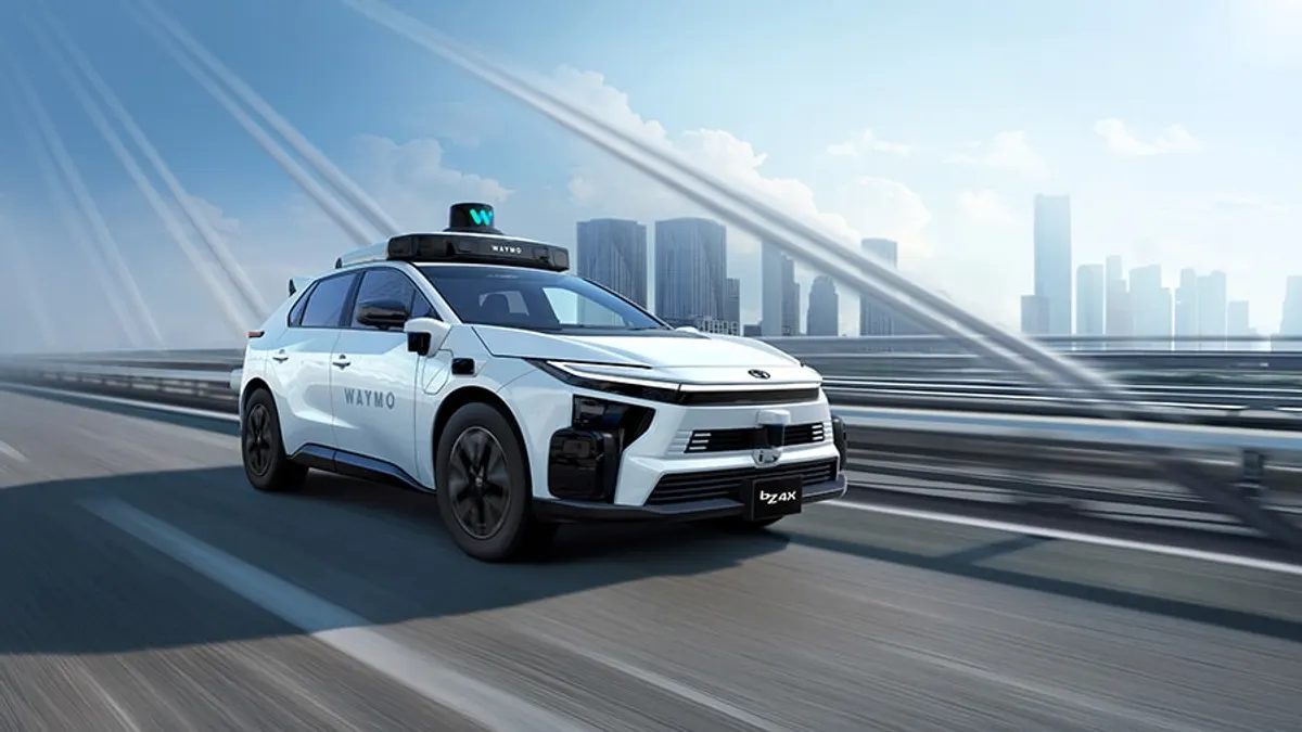 White Toyota bZ4X electric SUV with Waymo branding and roof-mounted autonomous driving sensors, shown from a front three-quarter angle on a cable-stayed bridge with a city skyline in the background.