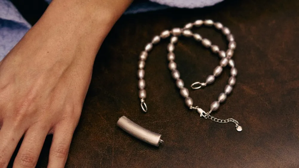 Nirva ambient AI pendant hardware capsule crafted from brushed metallic alloy beside a beaded jewelry necklace chain